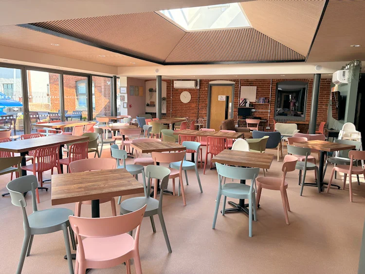 Inside The Canopy at Wymondham Baptist Church: wooden cafe tables with pink and grey chairs under a ceiling with a central skylight, glass doors along one side and the old brick wall of the church on the other